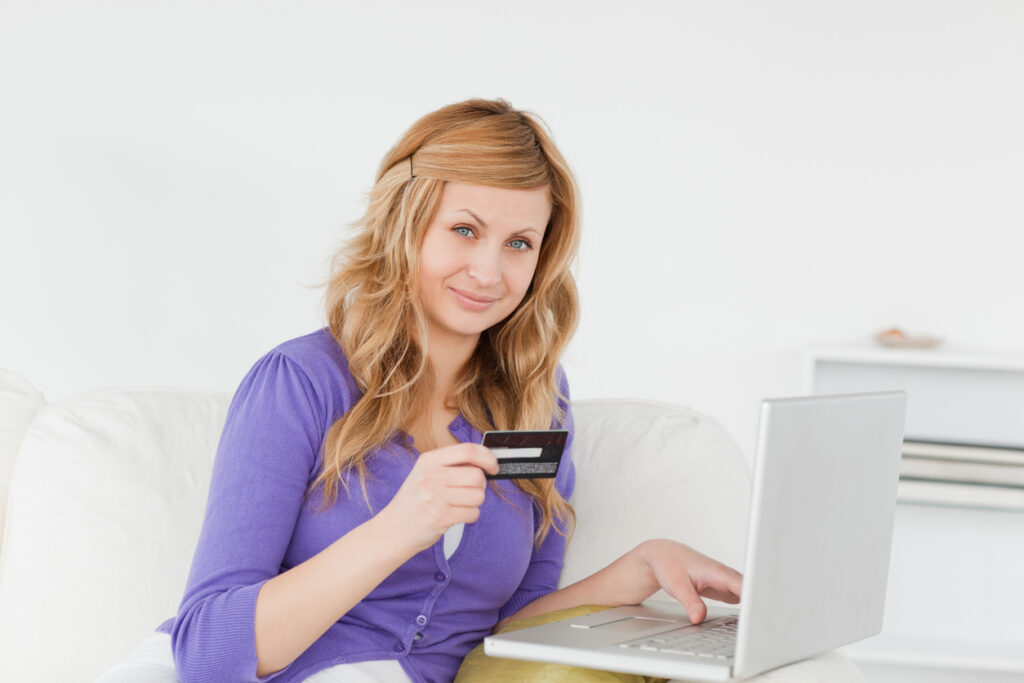 Mulher segurando cartão de crédito do Nubank Rewards utilizando notebook sentada no sofá..