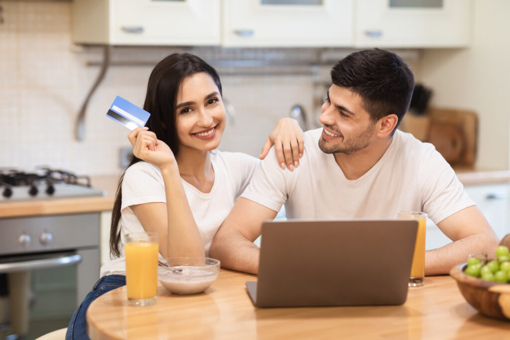 Transação online. Retrato de uma jovem encantada segurando um cartão de crédito, sentada com o pc em casa, abraçando o marido.