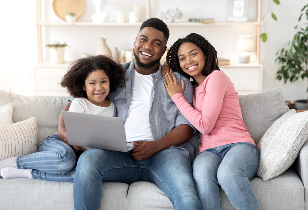 Família feliz sentada no sofá da sala, com o pai e a filha segurando o notebook fazendo compras online.