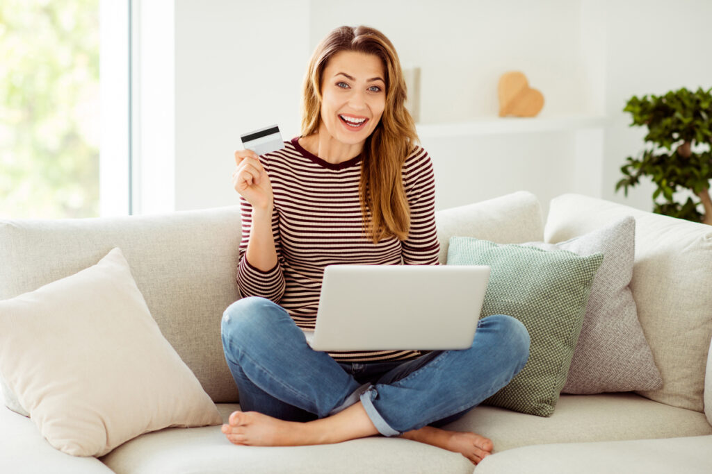 Mulher sorridente sentada no sofá da sua sala segurando um cartão de crédito em suas mãos e fazendo compra online no notebook.