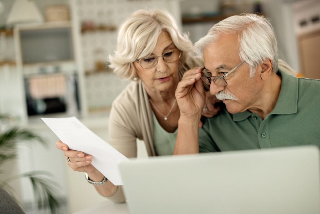 Casal de idosos conferindo o saldo de pontos da sua fatura de cartão de crédito. Casal sentado na mesa em frente ao notebook conferindo documentos.