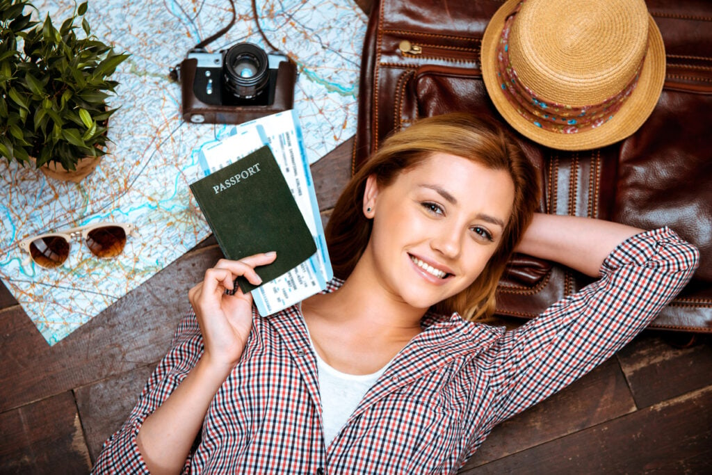 Linda menina loira deitada no chão de madeira. Mulher jovem sorrindo, segurando o passaporte e sua passagem aérea, e olhando para a câmera. Câmera vintage, chapéu e mapa estão no chão.