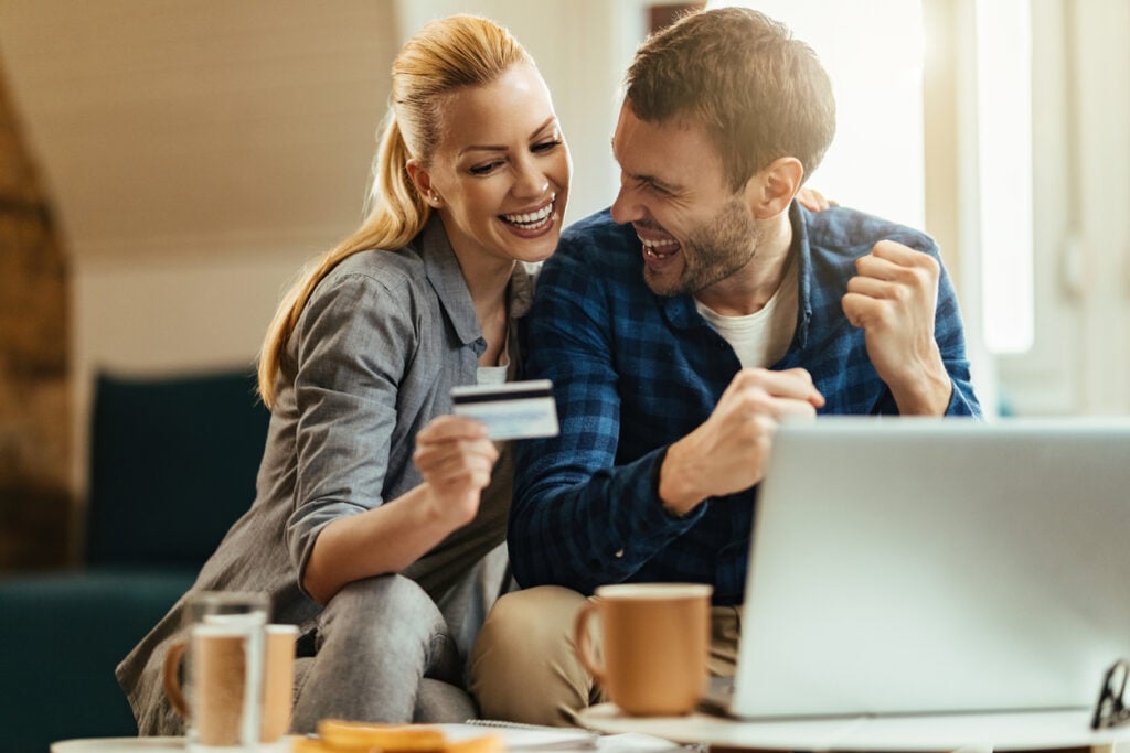 Casal feliz pois descobriram a hora ideal para vender milhas. Mulher segurando cartão de crédito na sala de estar com notebook em cima da mesa e seu marido comemorando.