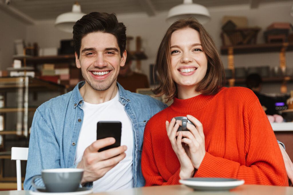 Casal sorridente sentados tomando um café, utilizando o celular para utilizar o App da Hot Milhas.